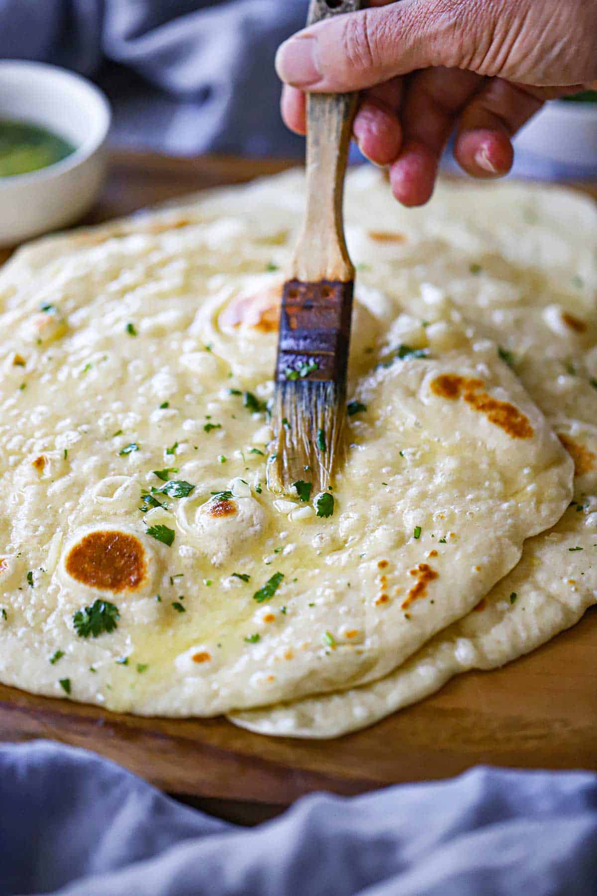 A person using a pastry brush to apply a melted garlic-cilantro butter over the top of a stack of homemade garlic naan on a wooden platter.