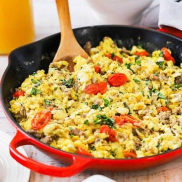 A close-up view of a large skillet with a red base filled with an Italian Skillet Scramble topped with blistered cherry tomatoes and chopped basil.