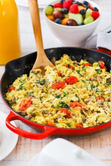 A straight-on view of a large skillet with a red base filled with an Italian Skillet Scramble with a wooden spoon inserted into the side of the eggs and a bowl of mixed fruit in the background.