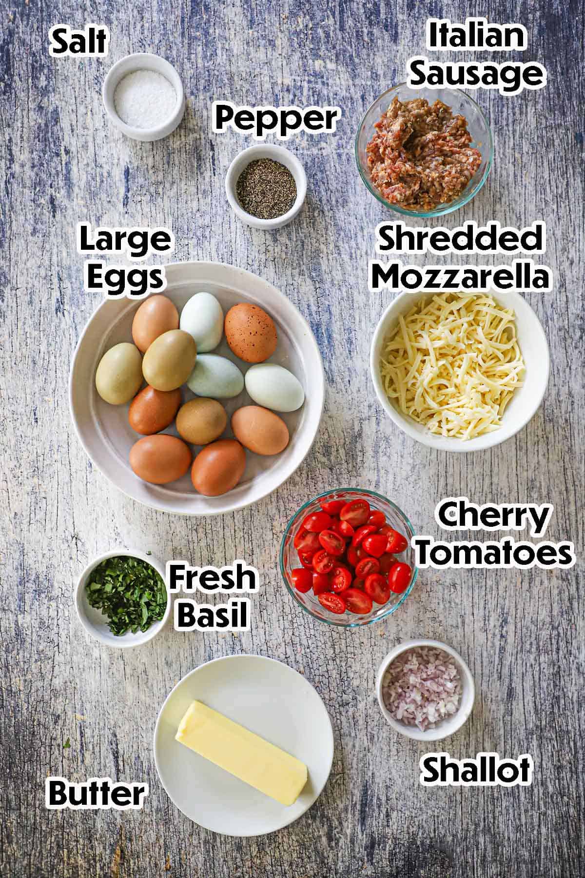 An arrangement of ingredients for Italian Skillet Scramble on a grey-wooden background including fresh eggs, Italian sausage, Mozzarella, cherry tomatoes, shallots, butter, basil, and seasonings.