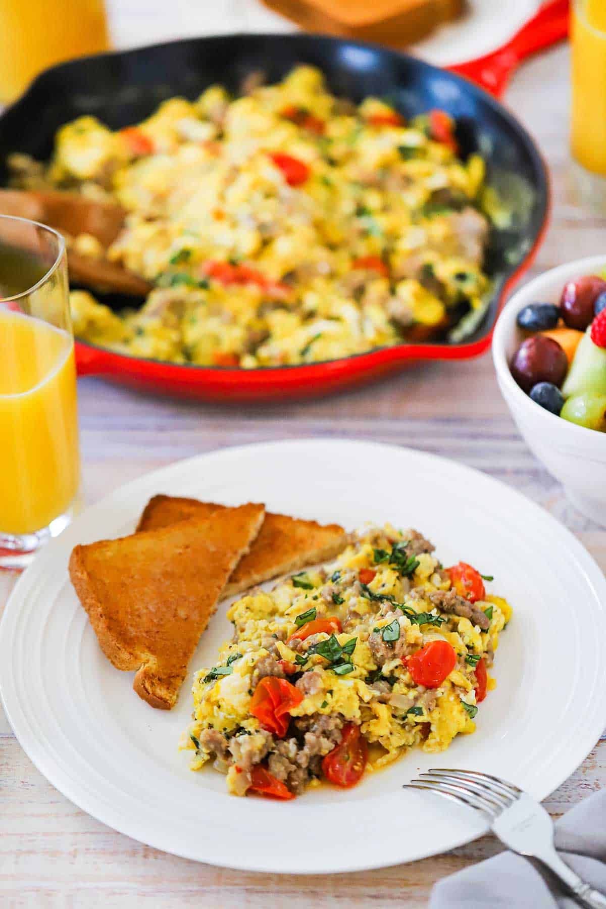 A white dinner plate filled with a serving of Italian Skillet Scramble next to two slices of toast with a skillet filled with the eggs in the background.