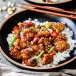 A staight-on view of a black shallow bowl filled with a serving of crispy kung pao cauliflower on top of steamed white rice garnished with strips of scallions and toasted sesame seeds.