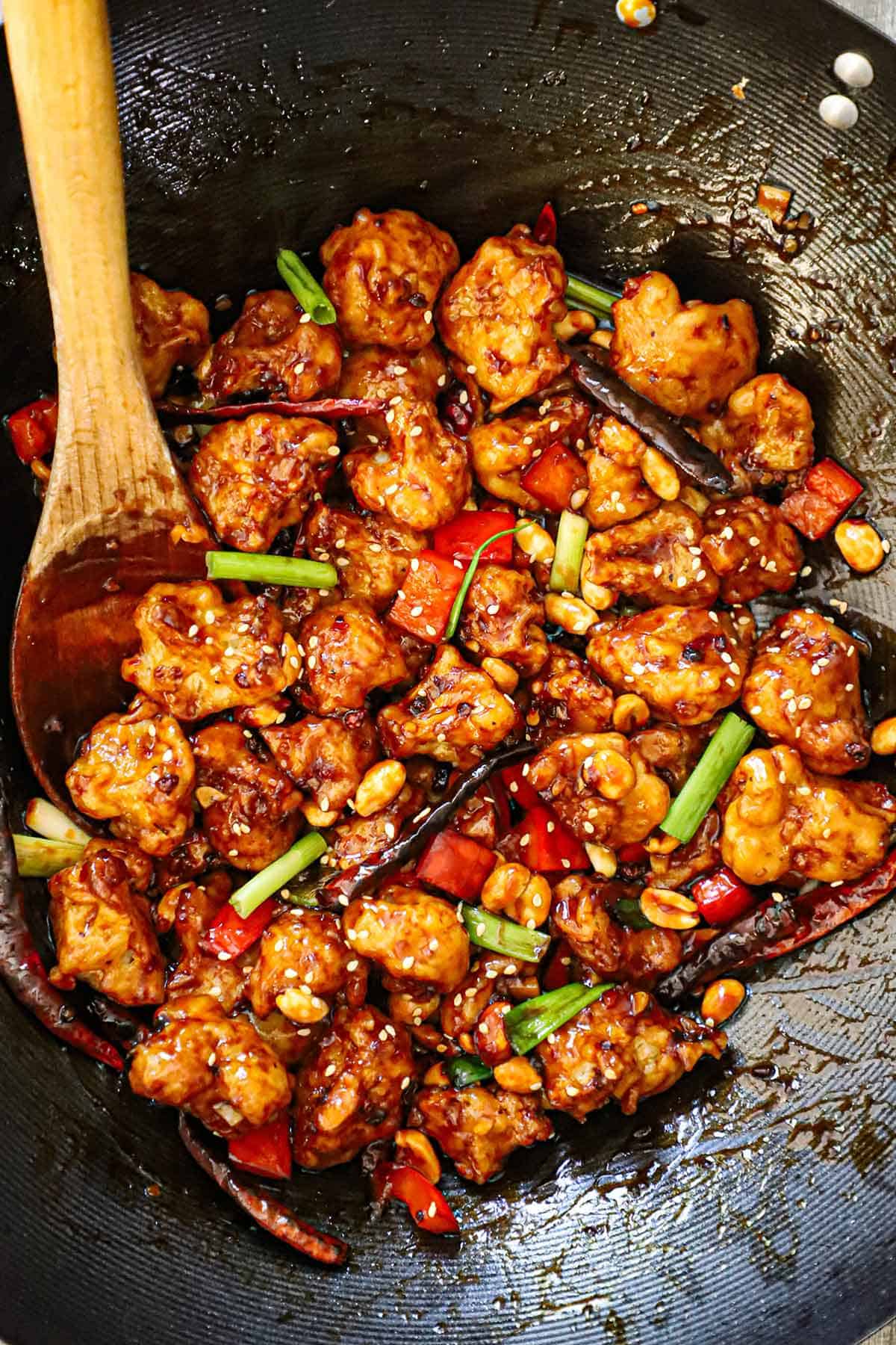 An overhead view of a black wok that is filled with freshly prepared crispy kung pao cauliflower topped with strips of scallions, dried red chiles, peanuts, and sesame seeds.