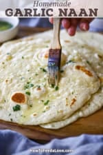 A person using a pastry brush to apply a melted garlic-cilantro butter over the top of a stack of homemade garlic naan on a wooden platter.
