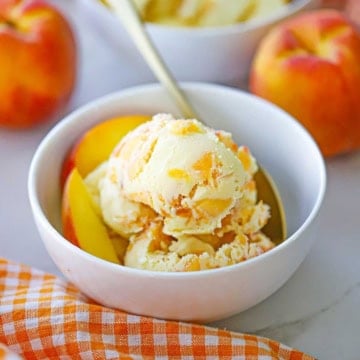 A white ice cream bowl filled with several scoops of homemade peach ice cream with a orange checkered napkin nearby.