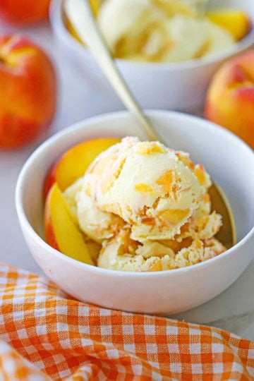 A straight-on view of several scoops of homemade peach ice cream with a couple of peach slices all in a white ice cream bowl.
