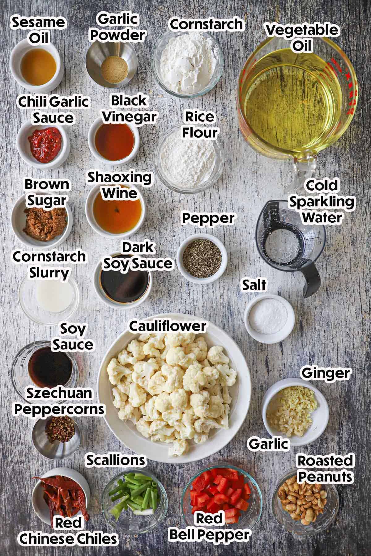 An arrangement of ingredients for crispy kung pao cauliflower on a grey wooden background including cauliflower florets, rice flour, cornstarch, ginger, garlic, peanuts, brown sugar, red bell pepper, scallions, red chiles, spices, and seasonings.