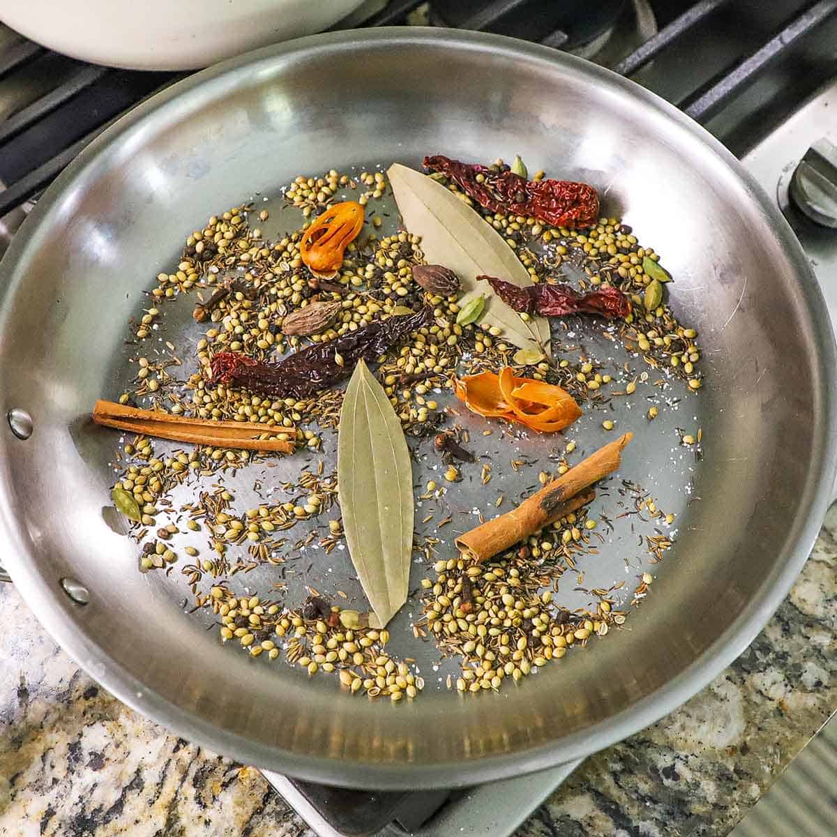 A large silver skillet on a gas stove containing bay leaves, cinnamon stick, and dried spices for biryani masala.