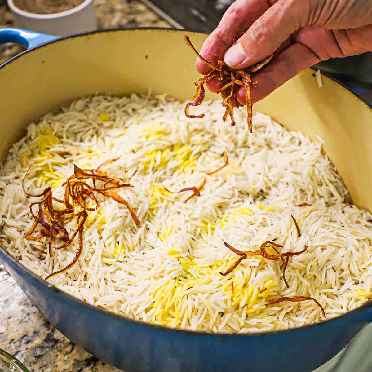 A person sprinkling thin strips of fried onions over the top of partially cooked basmati rice that is resting on uncooked marinated chicken in a Dutch oven.