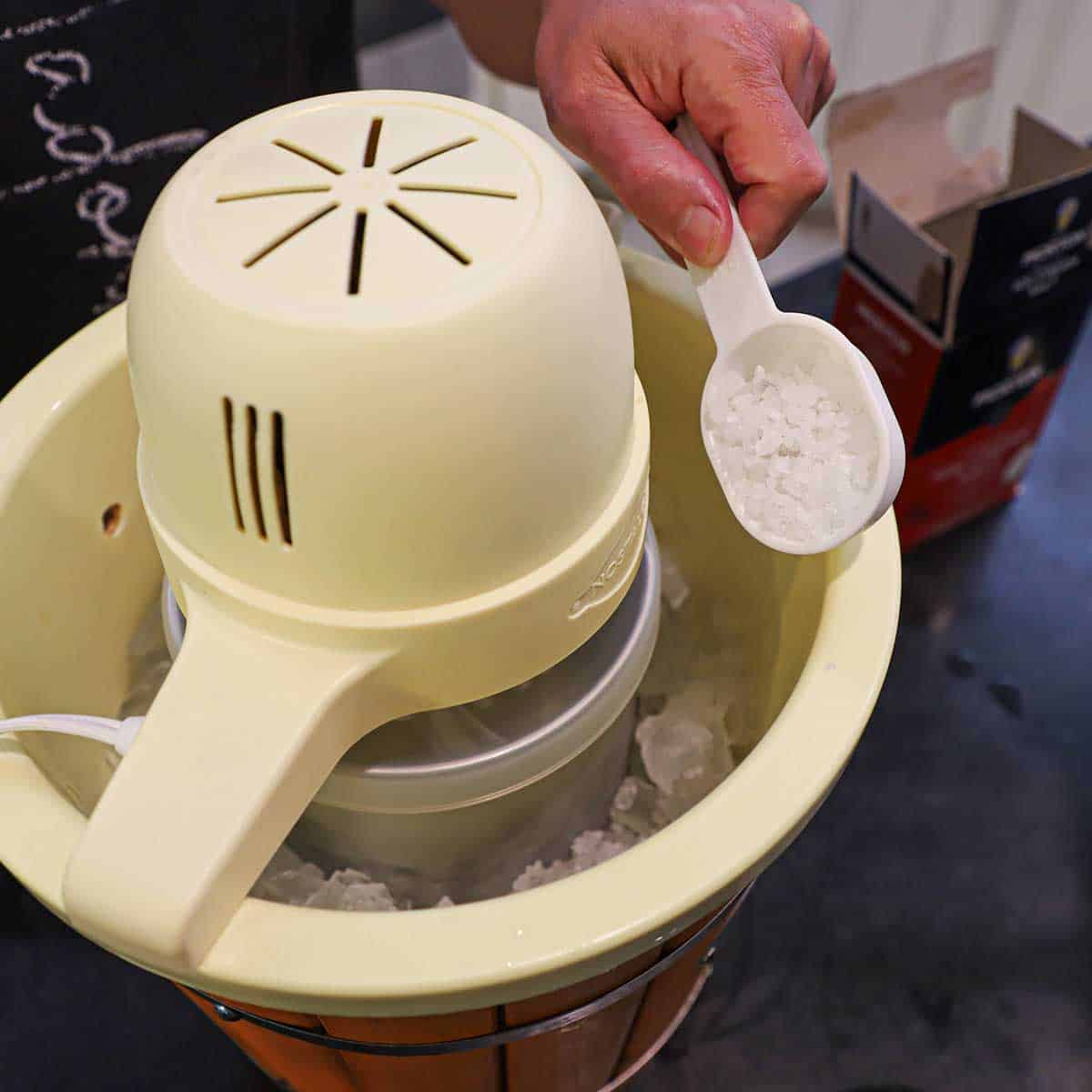 A person transferring rock salt from a small plastic measuring cup into an electric ice cream make that is partially filled with ice.