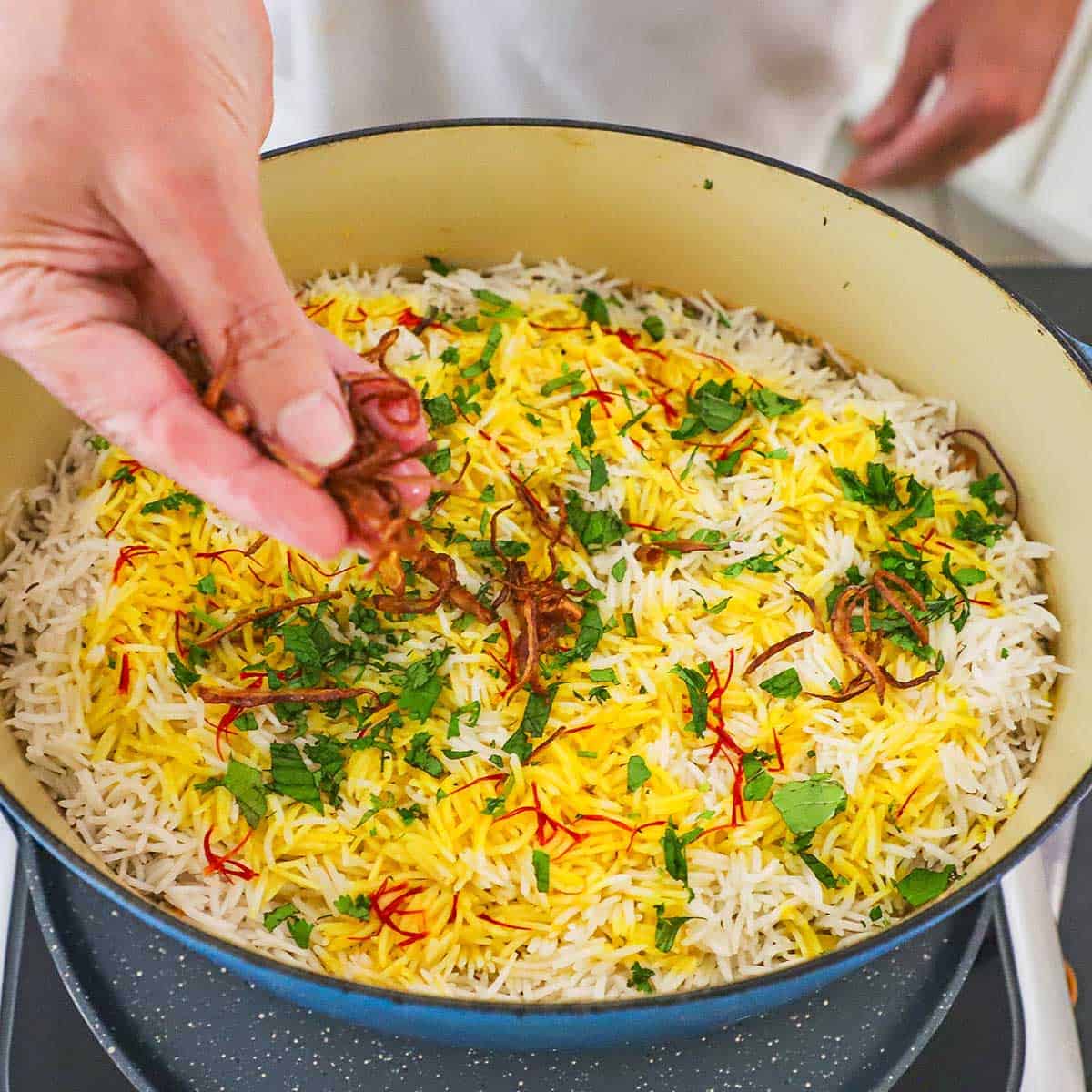 A person sprinkling fried onions over the top of uncooked Hyderabadi Chicken Biryani in a large Dutch oven.