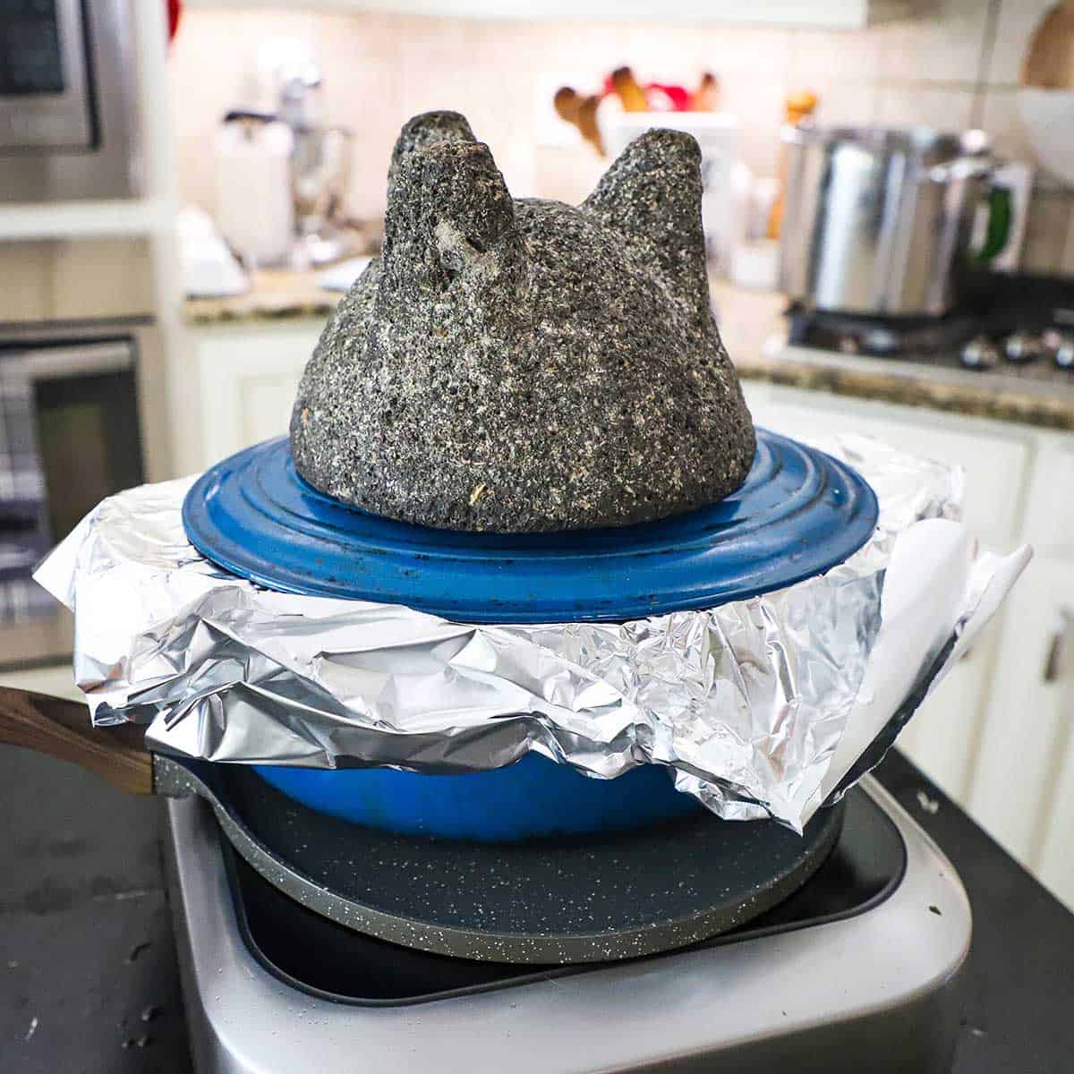 A molcajete turned upside down resting on the lid of an oval Dutch oven that is lined with foiled and is resting on a comal on a induction oven on a kitchen island.