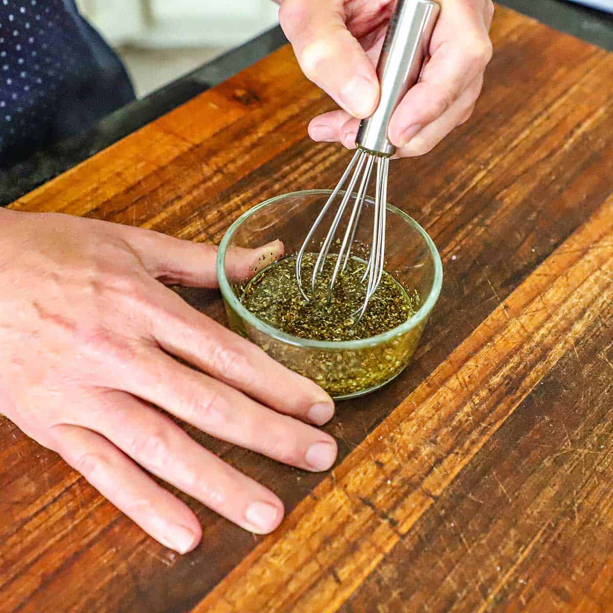 A person using a small whisk to combine olive oil with garlic and seasonings in a small glass bowl on a wooden cutting board.