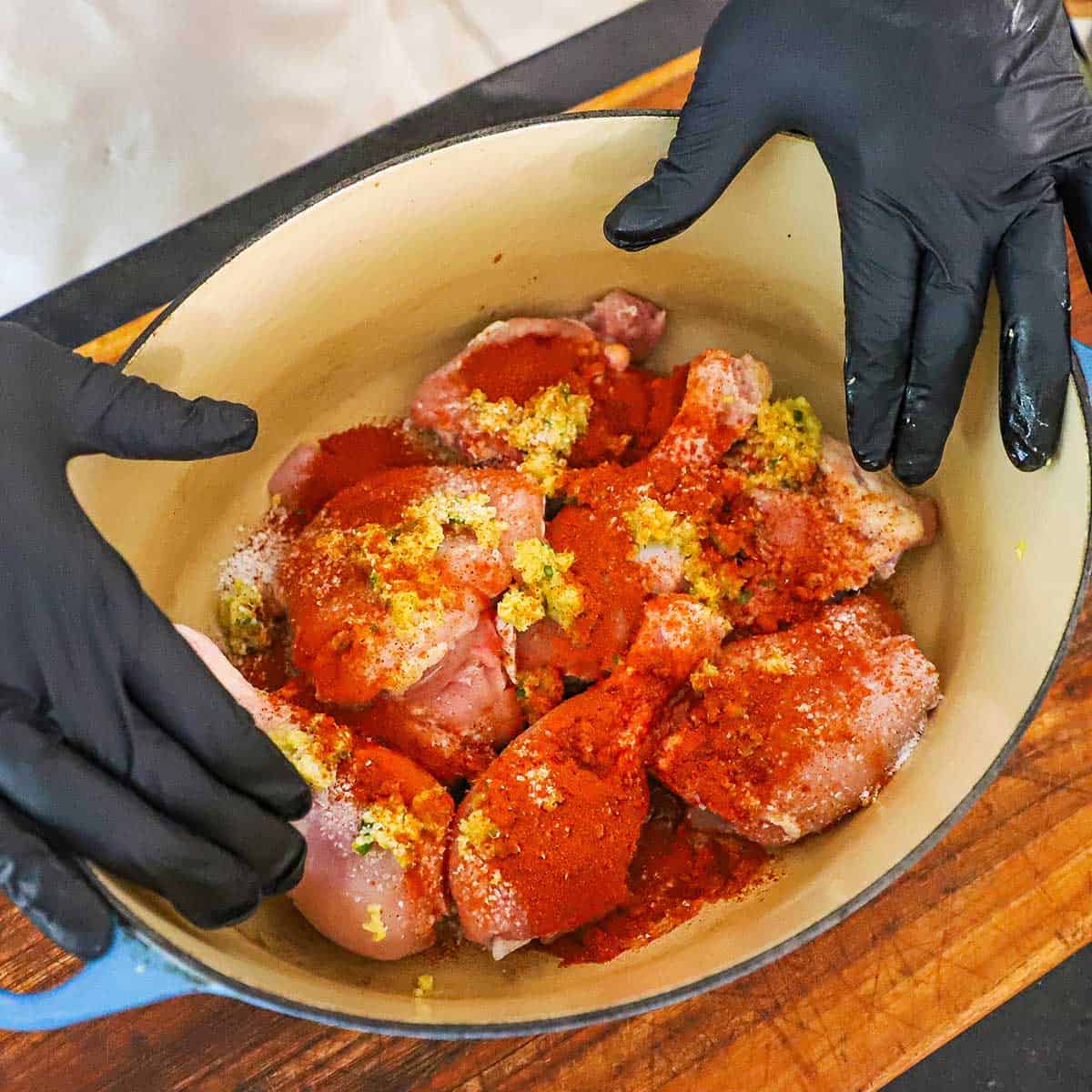 A person wearing black kitchen gloves preparing to rub chili powder, ginger-garlic paste, and lemon juice into chicken pieces in a large Dutch oven.