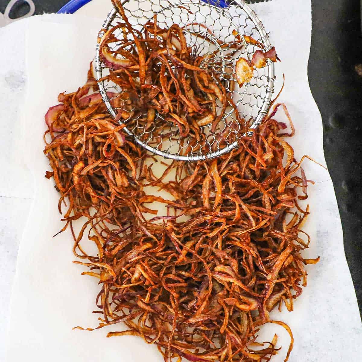 A person using a metal spider to place thin, fried onion strips onto a platter that is lined with paper towels.