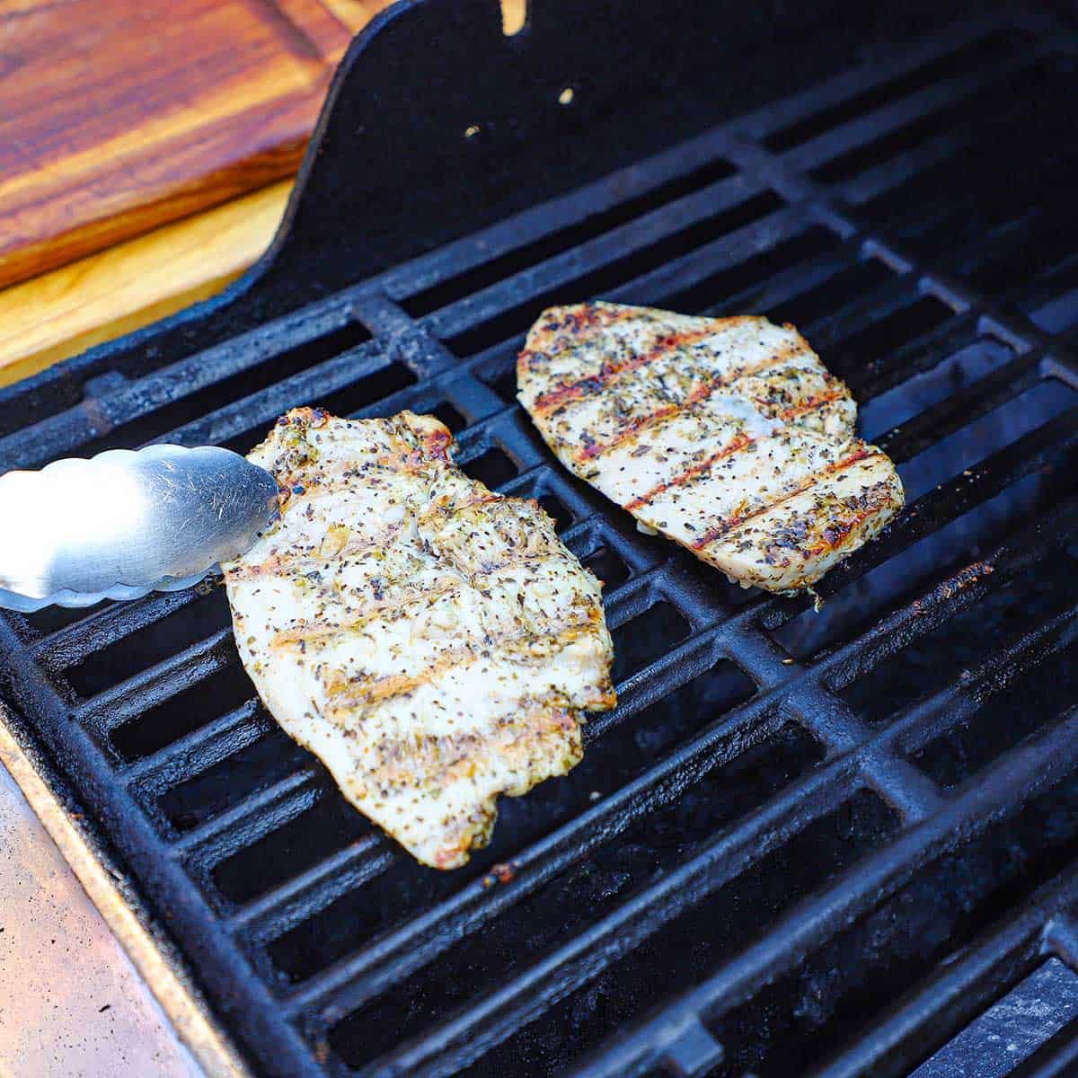 A person using a pair of metal tongs to turn grilled chicken cutlets that have been marinated in an herb marinade.