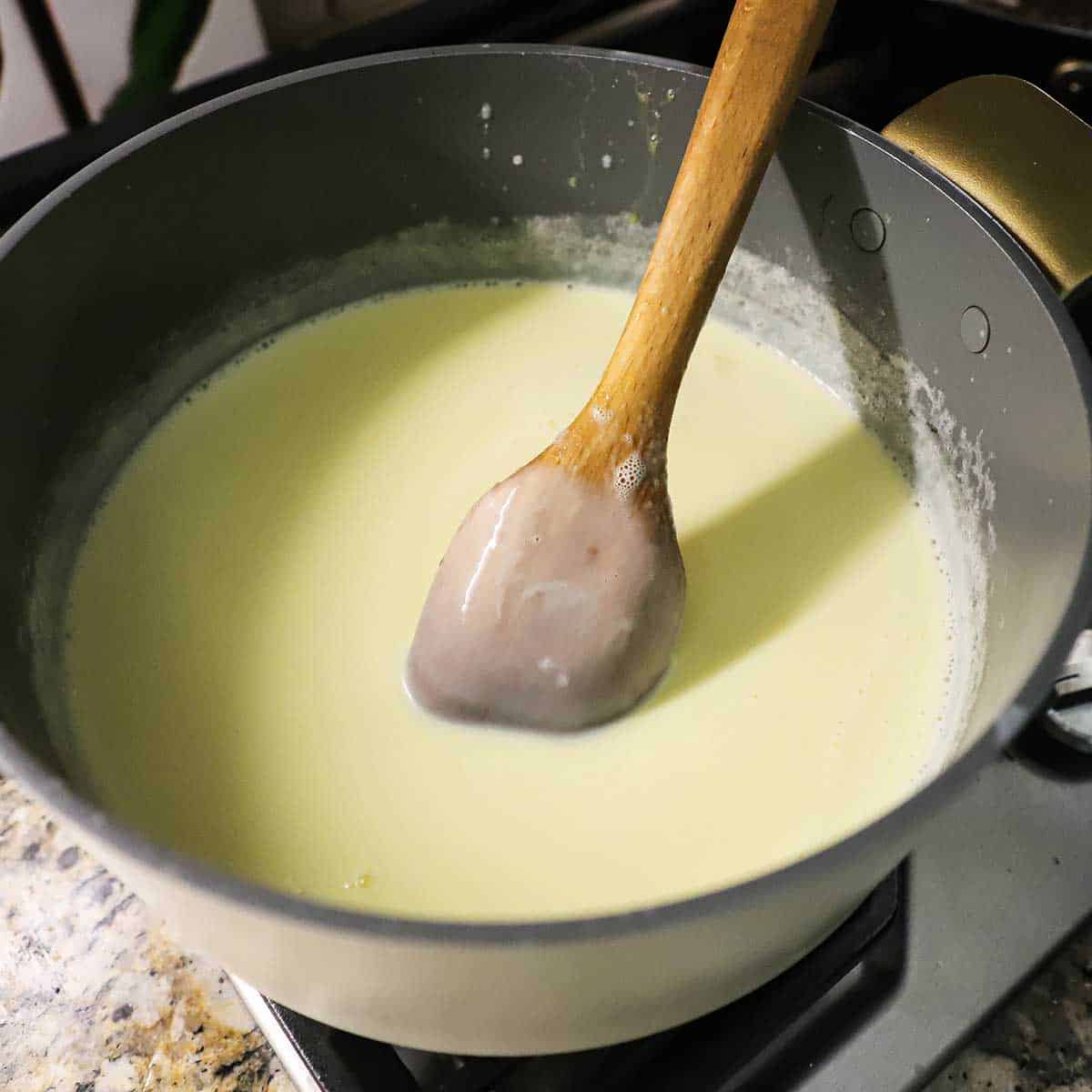 A person using a wooden spoon to stir a warm vanilla custard in a large pot on a gas stove.