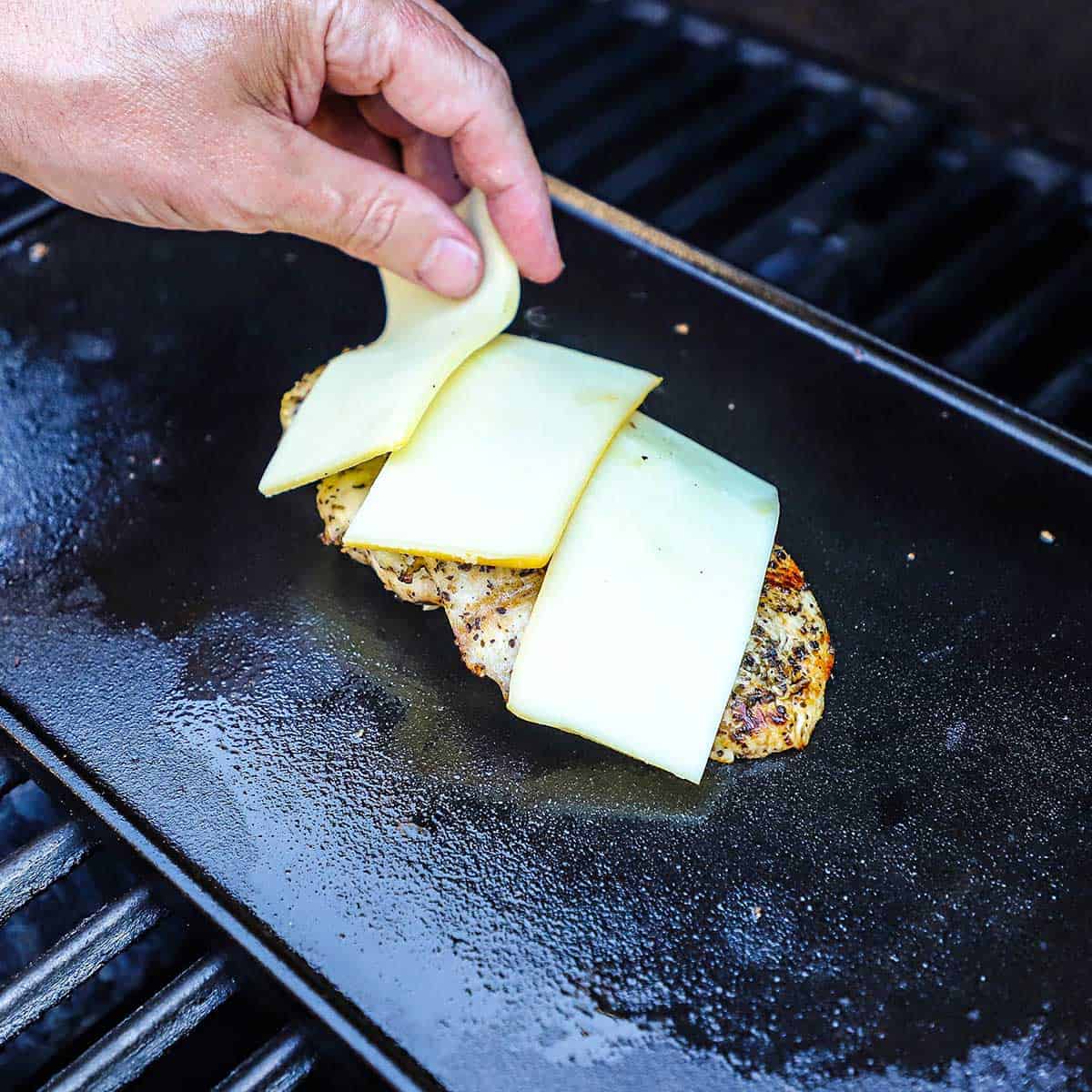 A person place strips of sliced smoked mozzarella over the top of a grilled chicken cutlet that is resting on a griddle on a gas grill.