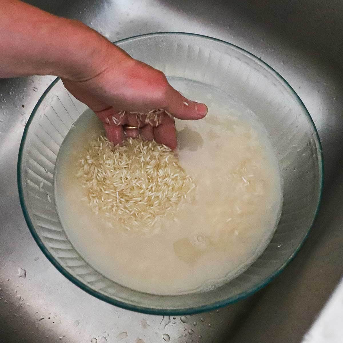 A person using one hand to swirl uncooked basmati rice in a water in a large glass bowl that is in the base of a kitchen sink.
