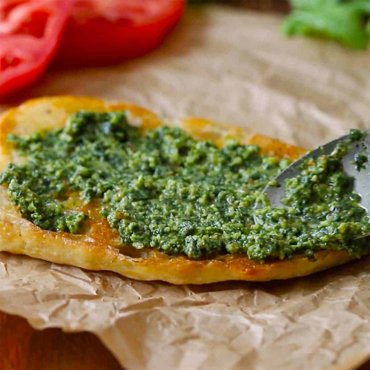A person using a spoon to spread homemade pesto sauce over a toasted bottom half of a ciabatta roll.