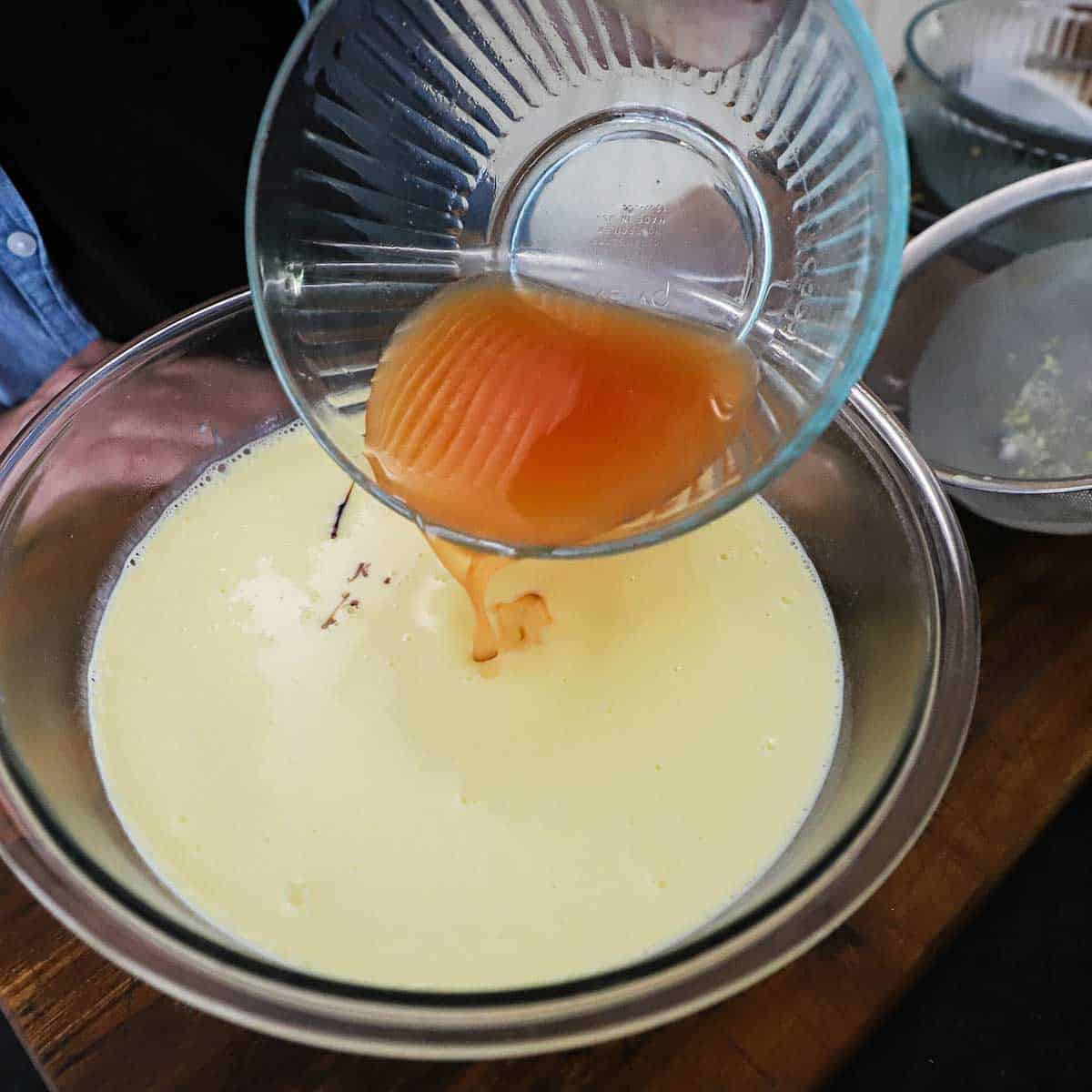 A person pouring the juice from macerated peaches from a glass bowl into another glass bowl filled with a warm vanilla custard.