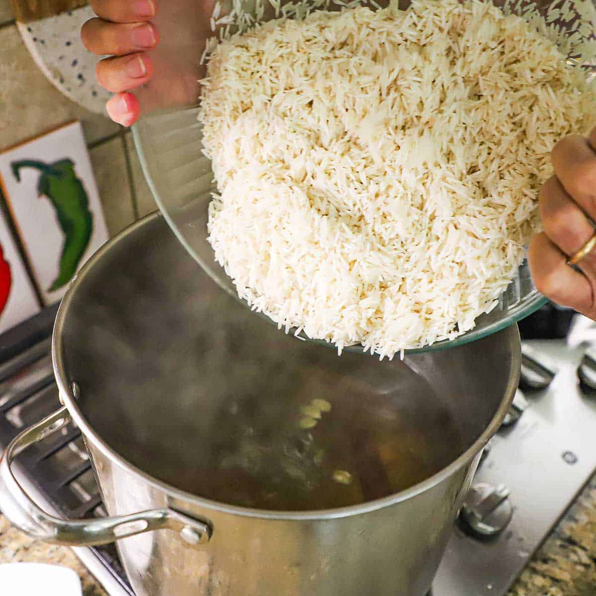 A person transferring soaked basmati rice from a large glass bowl into simmering, seasoned water in a large stock pot on a gas stove.