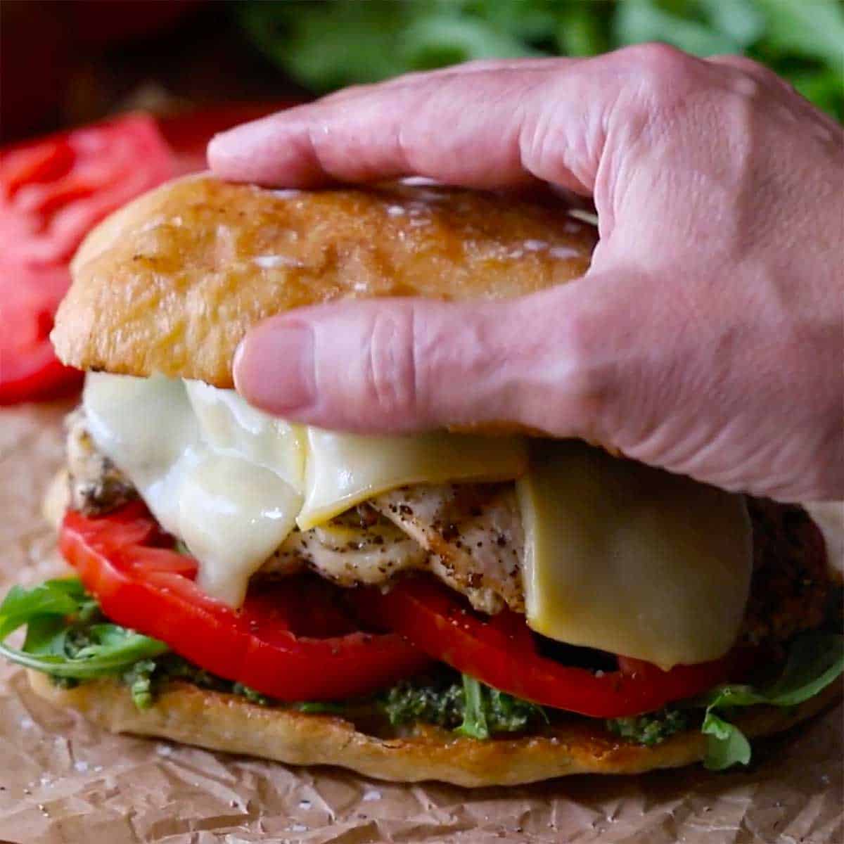 A person placing the top half of a toasted ciabatta roll onto the top of a grilled pesto chicken sandwich with smoked mozzarella.