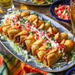 A close-up view of a platter filled with a line of crispy beef flautas topped with pico de gallo, crumbled Cotija cheese, and chopped cilantro.