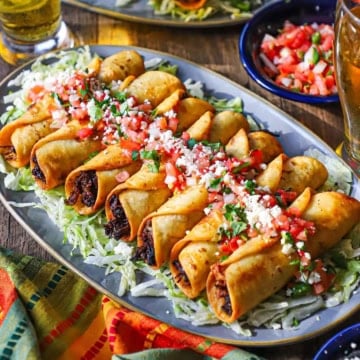 A close-up view of a platter filled with a line of crispy beef flautas topped with pico de gallo, crumbled Cotija cheese, and chopped cilantro.