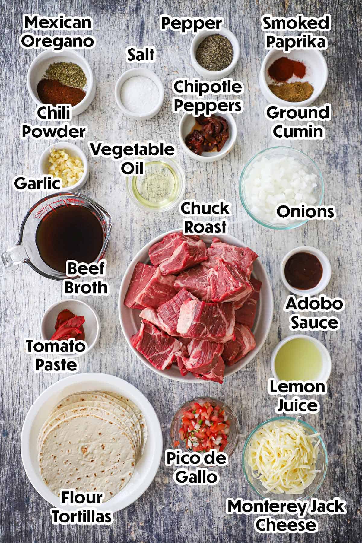 An arrangement of ingredients for crispy beef flautas on a grey wooden background including chuck roast, onions, garlic, oil, tomato paste, beef broth, lemon juice, chipotle peppers, seasonings, tortillas, and cheese.