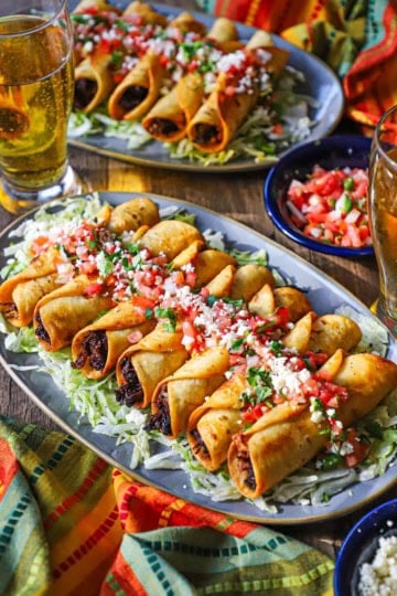 A straight-on view of two platters both filled with a line of crispy beef flautas topped with pico de gallo, crumbled Cotija cheese, and chopped cilantro.