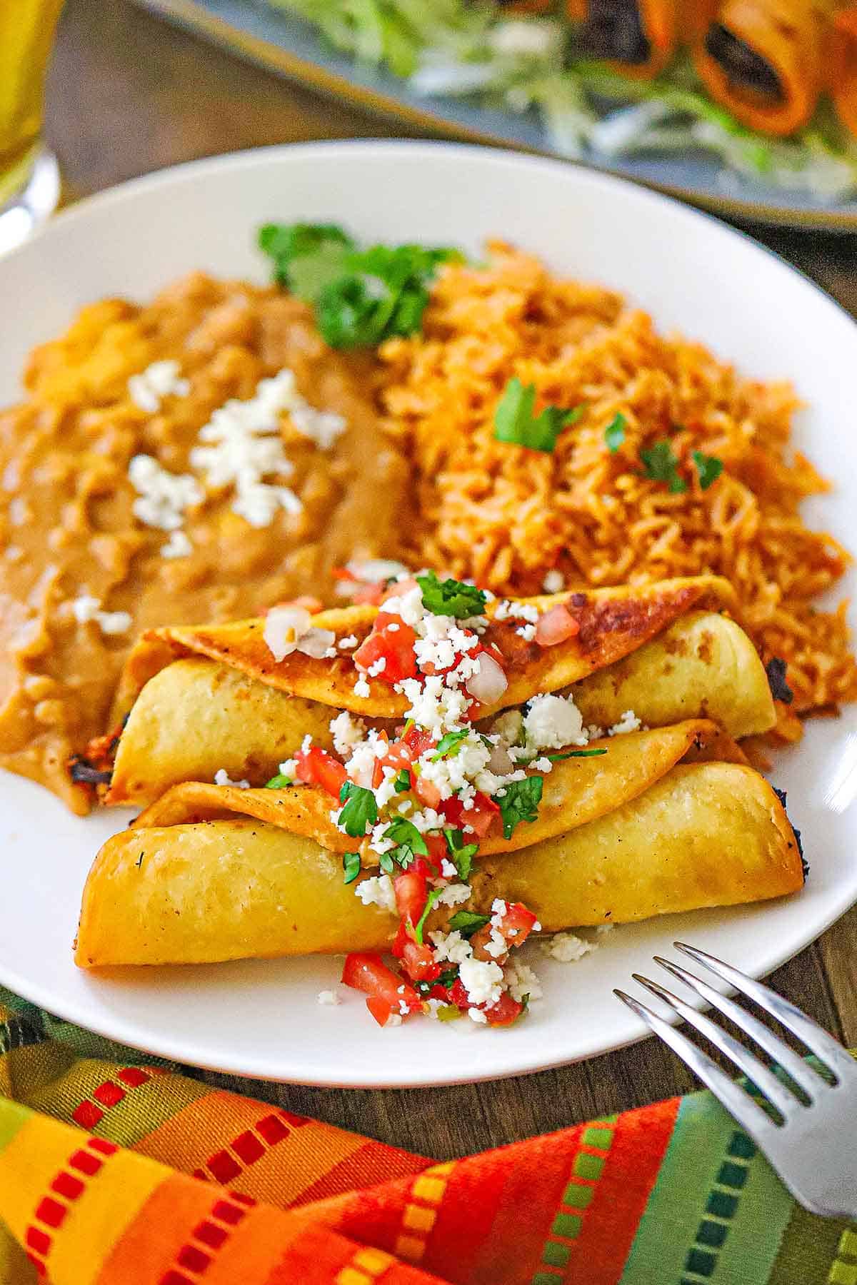 A straight-on view of a white dinner plate filled with a serving of two side-by-side crispy beef flautas next to a mound of Mexican rice and refried beans.