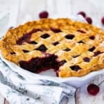 A close-up view of a homemade cherry pie in a white pie dish with a pasty lattice topping and a slice of the pie missing, revealing the cherry filling.