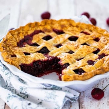 A close-up view of a homemade cherry pie in a white pie dish with a pasty lattice topping and a slice of the pie missing, revealing the cherry filling.