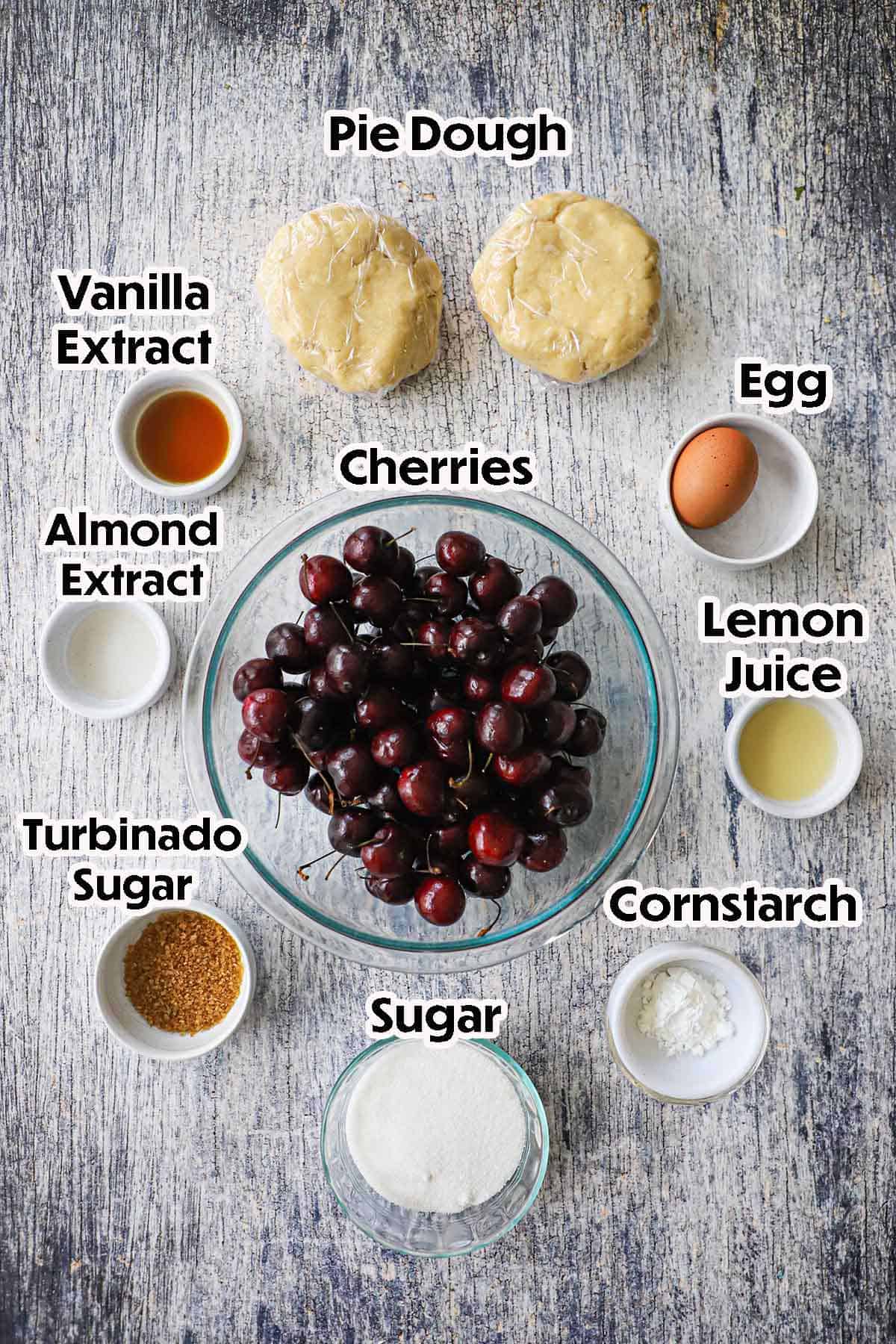 An arrangement of ingredients for homemade cherry pie on a grey wooden background including whole fresh cherries, lemon juice, sugar, cornstarch, an egg, vanilla extract, almond extract, and two discs of pie dough.