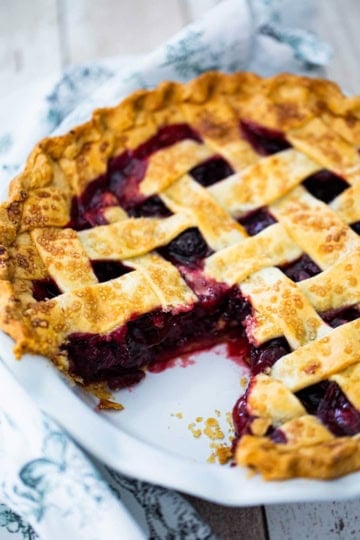 A straight-on view of a homemade cherry pie in a white pie dish with a pasty lattice topping and a slice of the pie missing, revealing the cherry filling.