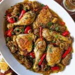 An overhead, close-up view of a large oval platter filled with Crispy Chicken Scarpariello consisting of seared chicken legs and thighs with chunks of Italian sausage all resting in a bell pepper and cherry pepper brown sauce.