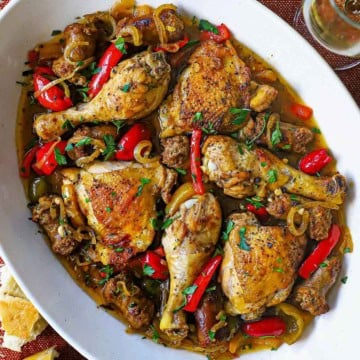 An overhead, close-up view of a large oval platter filled with Crispy Chicken Scarpariello consisting of seared chicken legs and thighs with chunks of Italian sausage all resting in a bell pepper and cherry pepper brown sauce.