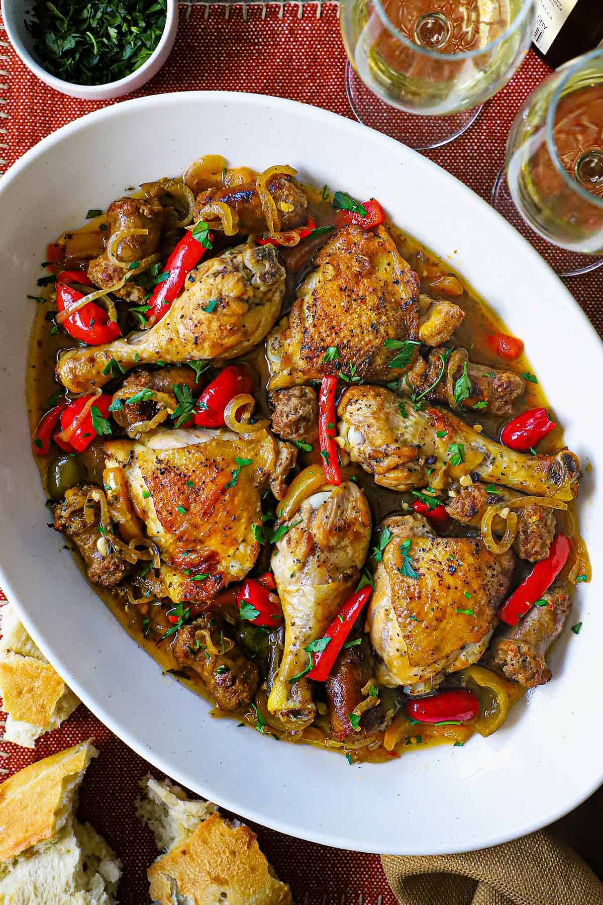An overhead view of a large oval platter filled with Crispy Chicken Scarpariello consisting of seared chicken legs and thighs with chunks of Italian sausage all resting in a bell pepper and cherry pepper brown sauce.