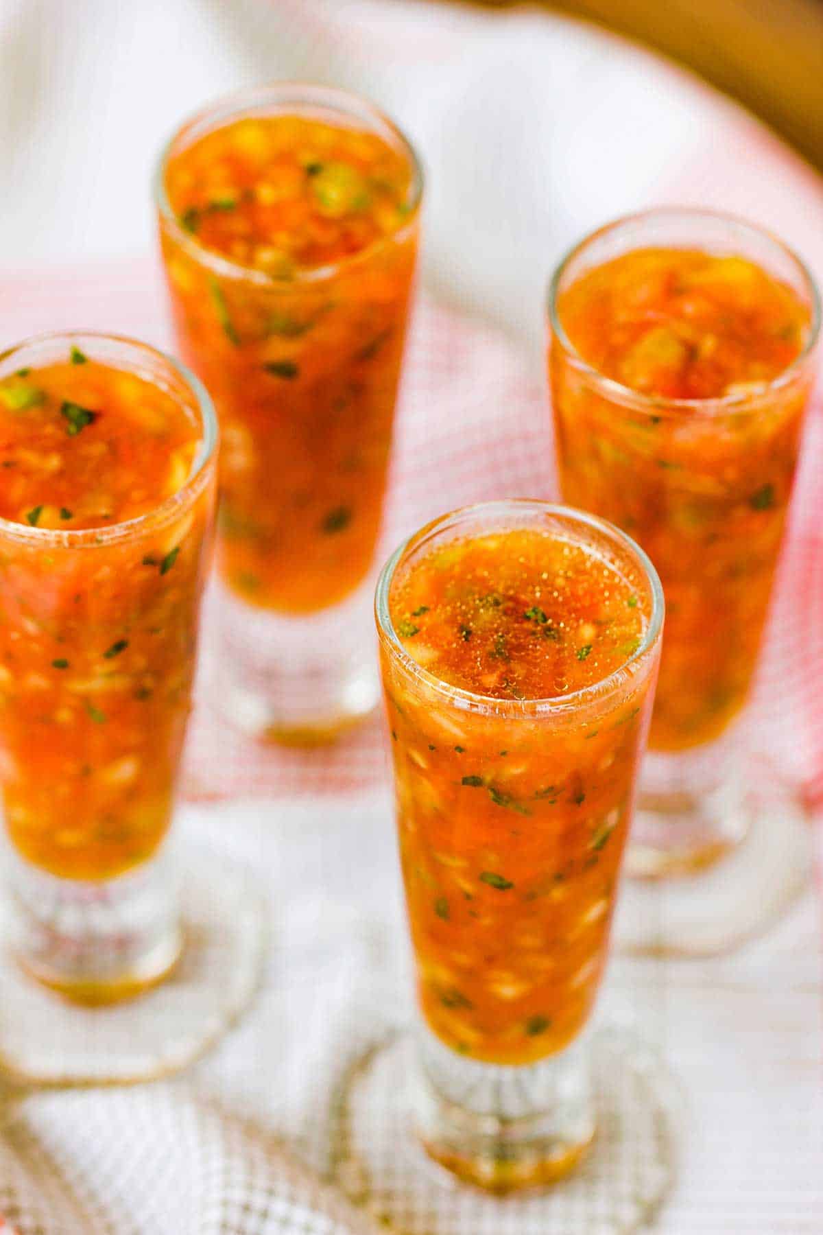 A close-up view of four tall, skinny clear shot glasses filled with classic gazpacho on a red and white checkered linen.