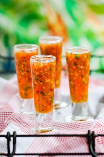 A close-up view of four tall, skinny clear shot glasses filled with classic gazpacho on a red and white checkered linen.