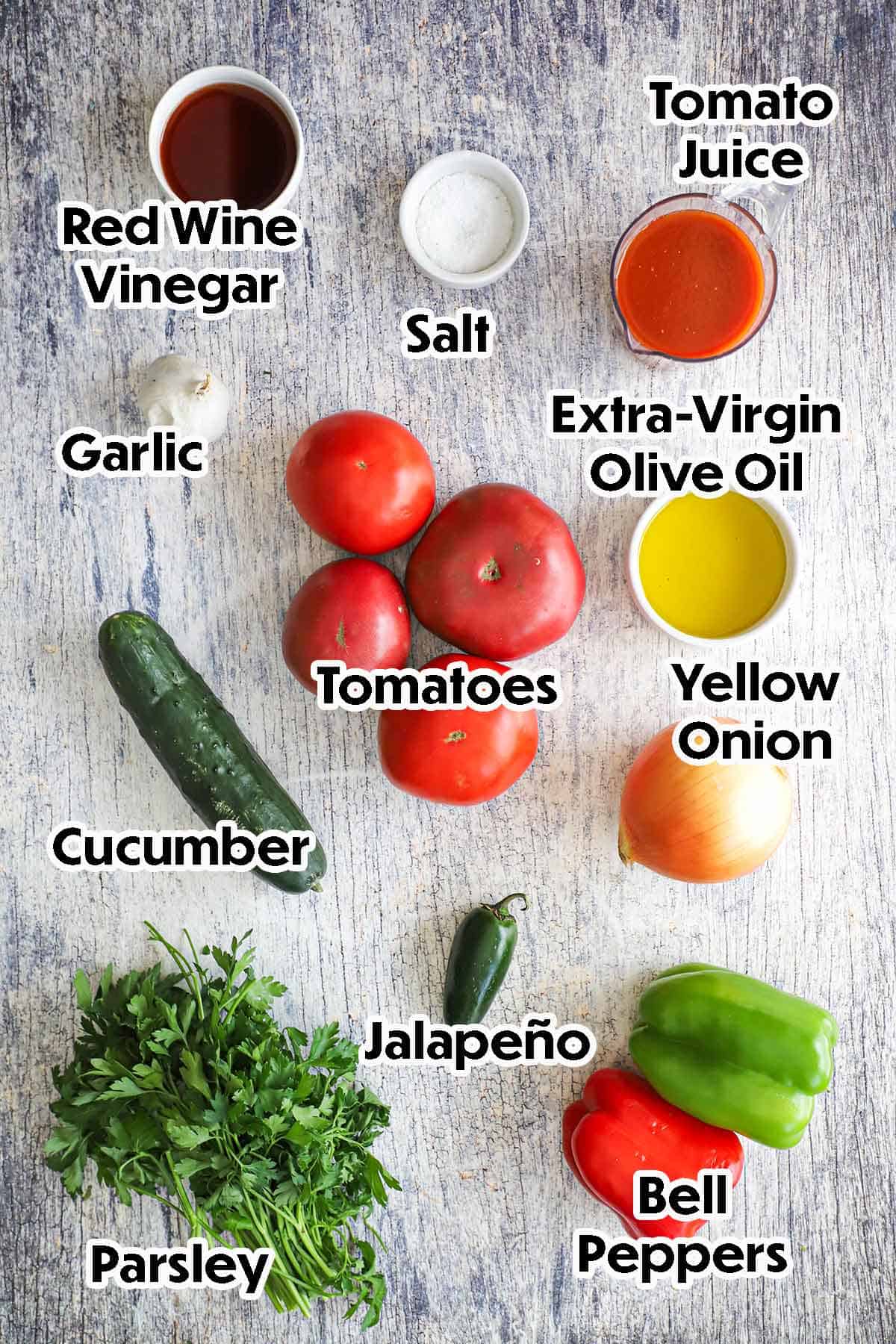An arrangement of ingredient for classic gazpacho on a grey wooden background including whole tomatoes, a cucumber, yellow onion, bell peppers, jalapeño, garlic, parsley, tomato juice, vinegar, oil, and salt.
