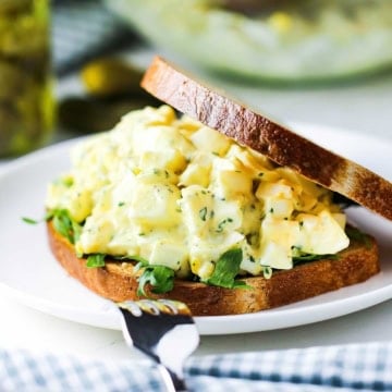 A close-up view a best-ever egg salad sandwich with toasted whole wheat sliced bread, egg salad, and arugula on a white plate.