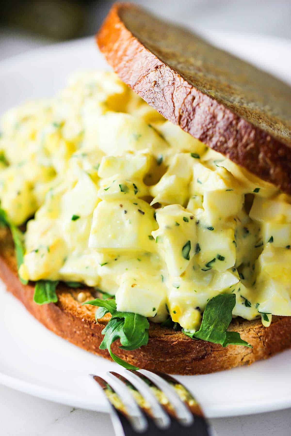 A close-up view a best-ever egg salad sandwich with toasted whole wheat sliced bread, egg salad, and arugula on a white plate.
