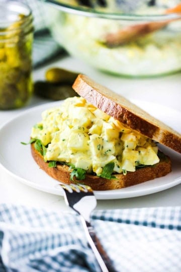 A straight-on view a best-ever egg salad sandwich with toasted whole wheat sliced bread, egg salad, and arugula on a white plate next to a jar filled with dill pickles.