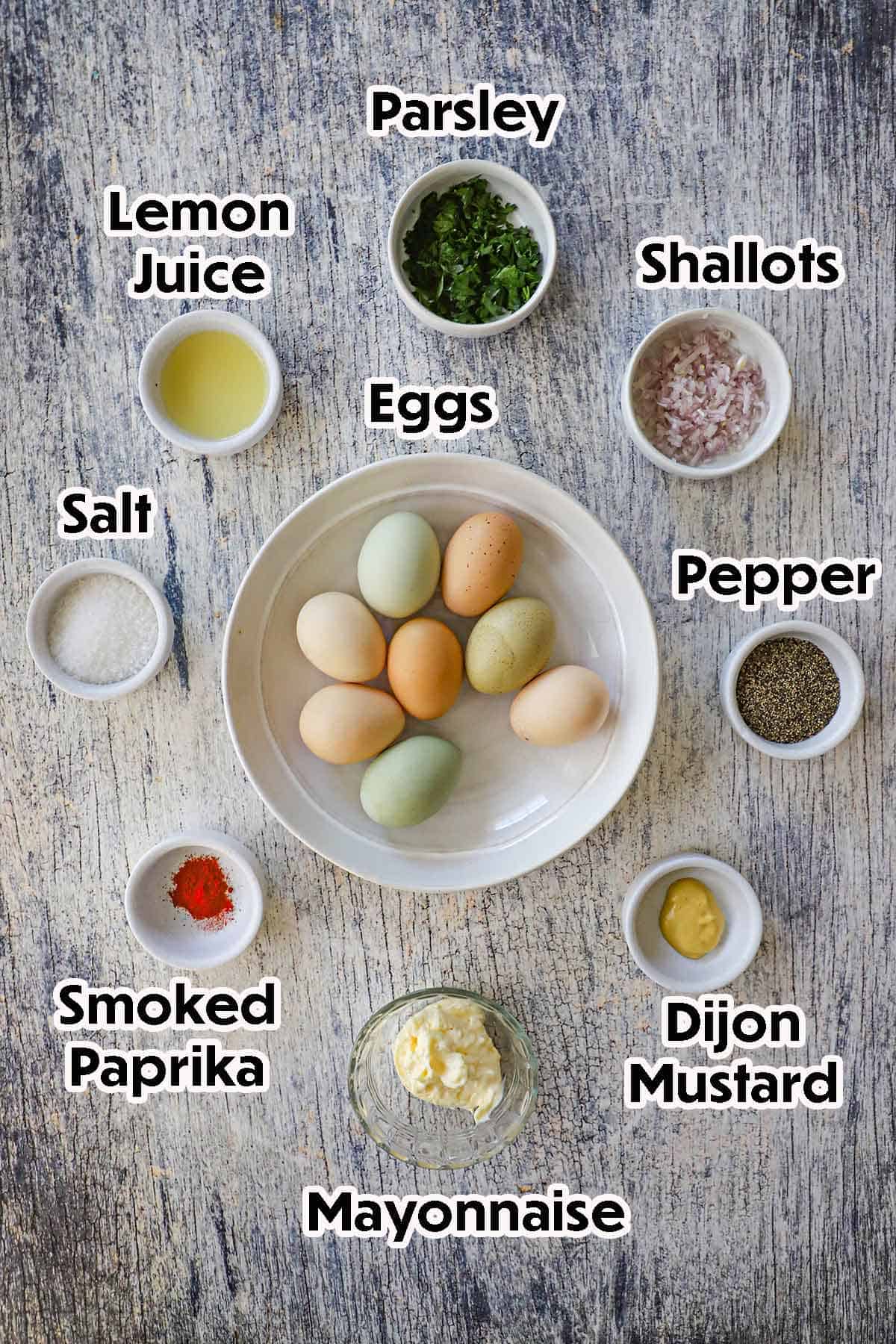 An arrangement of ingredients for best-ever egg salad sandwich on a grey wooden background including fresh eggs, mayonnaise, mustard, shallots, lemon juice, Parsley, and seasonings.