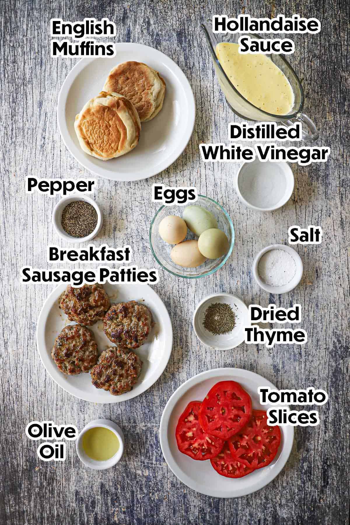 An arrangement of ingredients for Eggs Blackstone with Sausage on a grey wooden background including eggs, sausage patties, English muffins, tomato slices, hollandaise sauce, vinegar, olive oil, and seasonings.