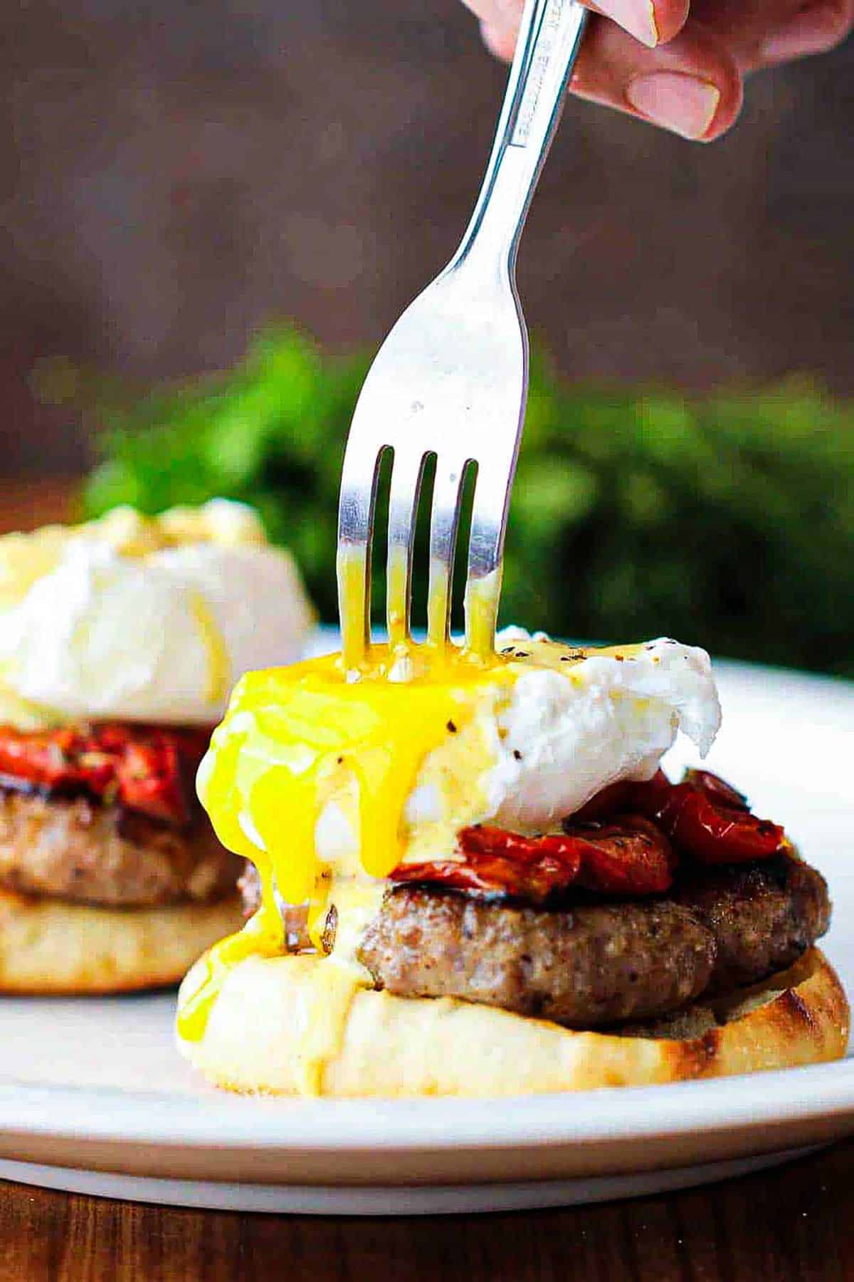 A person using a fork to plunge into a break open the yolk from a poached egg sitting on top of a roasted tomato slice resting on a sausage patty and toasted English muffin.