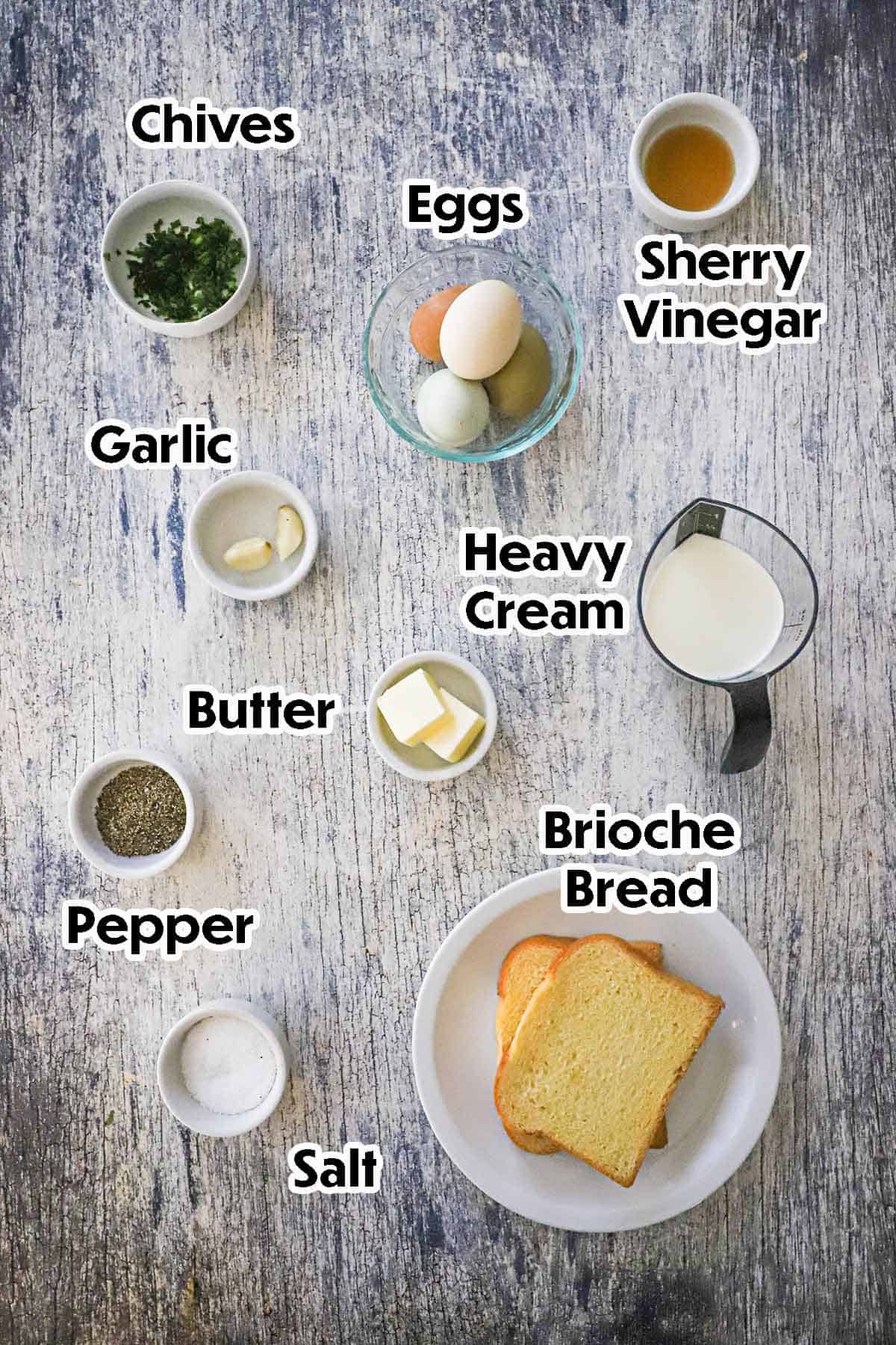 An arrangement of ingredients for French Baked Eggs with Cream on a grey wooden background including brioche bread, eggs, cream, butter, garlic vinegar, chives, and seasonings.