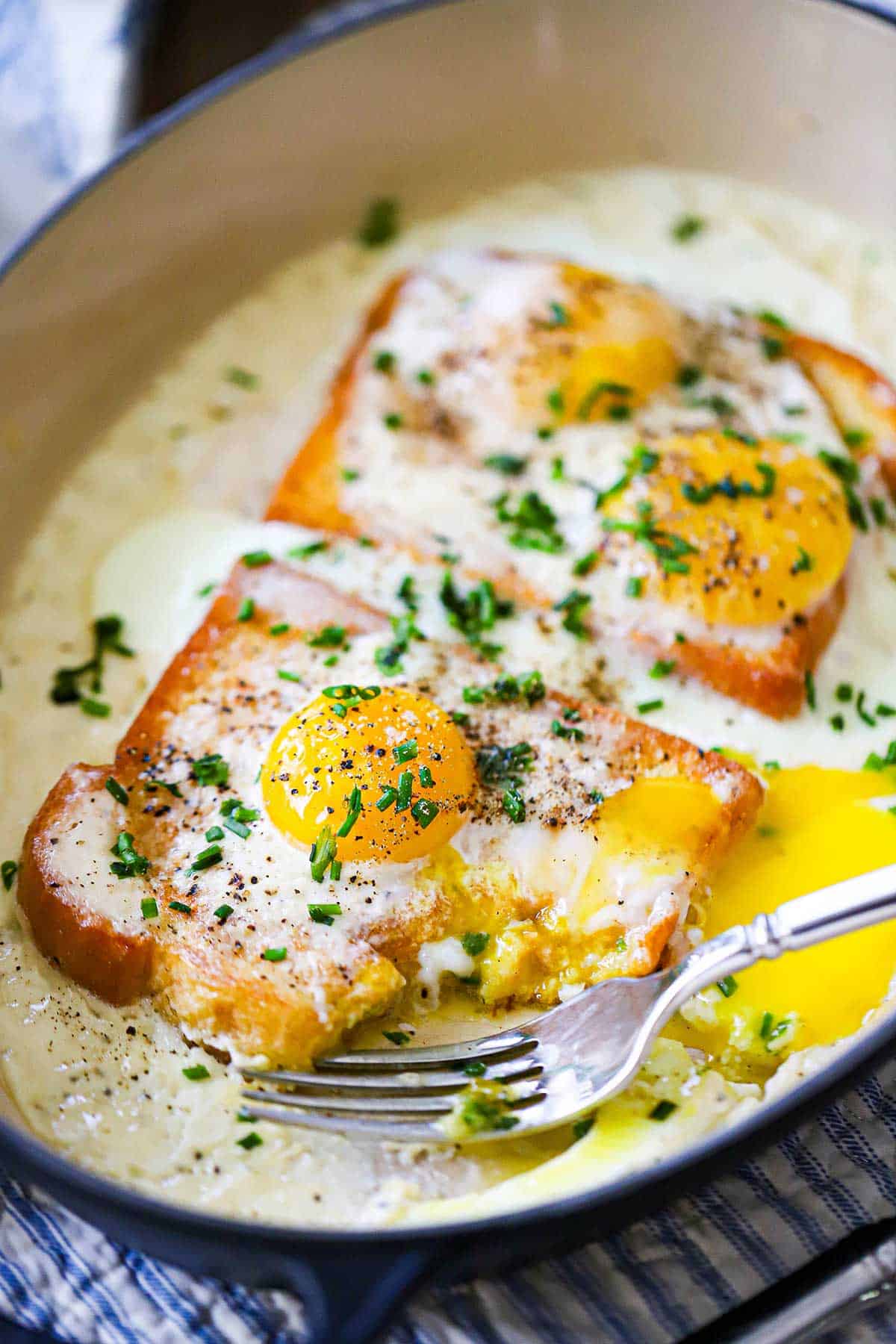 A close-up view of two French baked eggs in cream in an oval baking dish with part of the toast missing with egg yolk running throughout the dish.